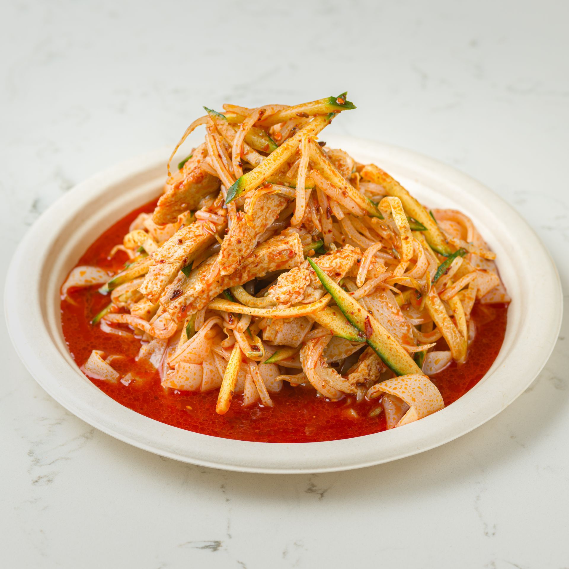 Spicy shredded salad with red sauce on a white plate, topped with herbs and chili flakes.