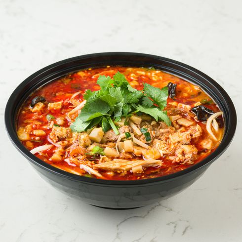 Bowl of spicy red soup topped with herbs and sliced vegetables on a white table