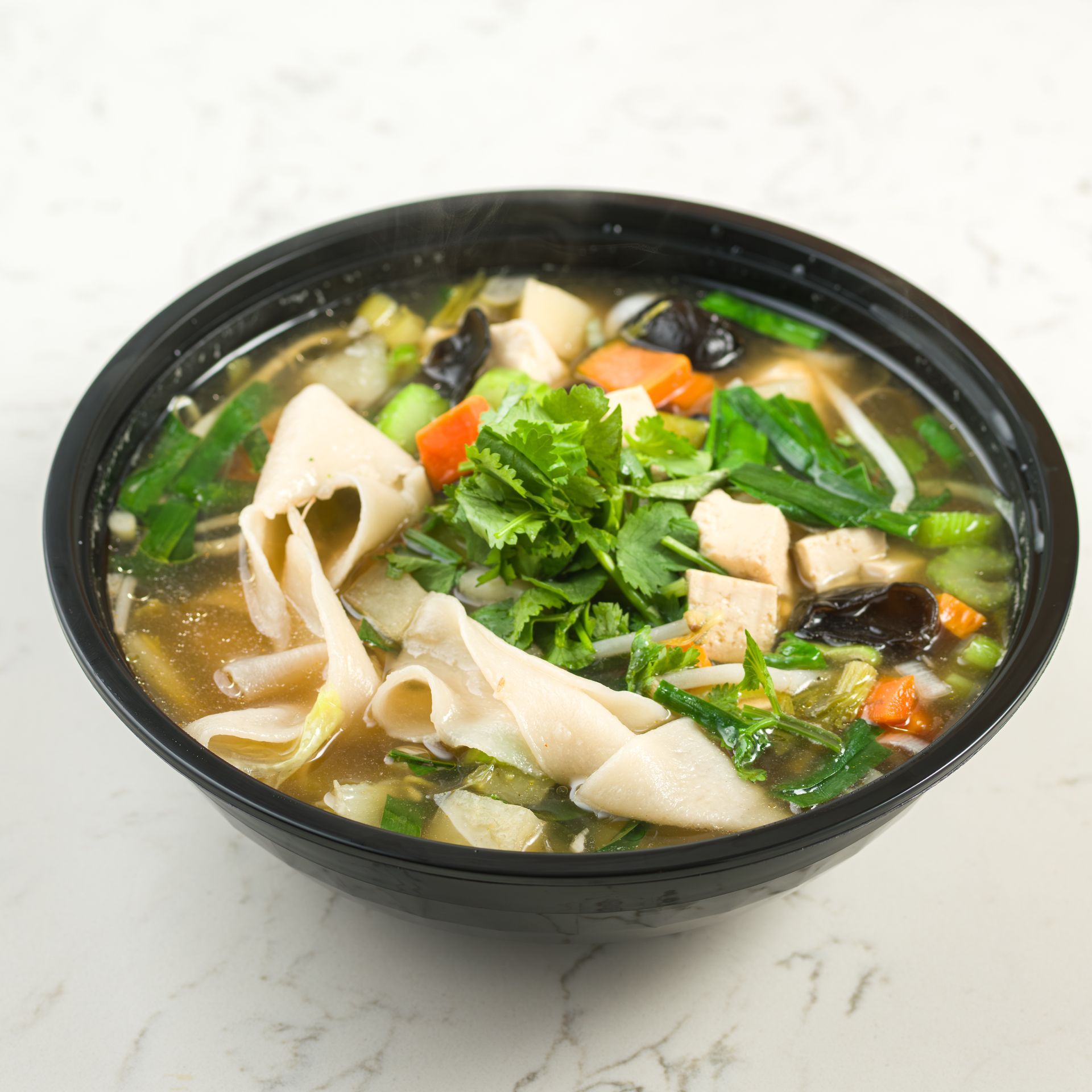 Black bowl of vegetable soup with greens, tofu, and noodles on a white surface