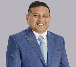 Romulo (Rom) A. Armas, Jr., man smiling wearing a white shirt, blue suit jacket, and light blue tie.