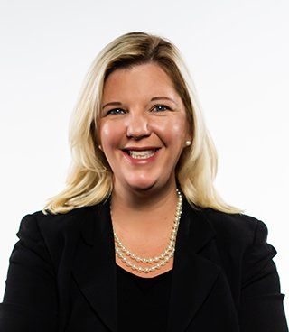Ashley Lovelace. Blonde woman, smiling, wearing a black top, black blazer, and pearl necklace.