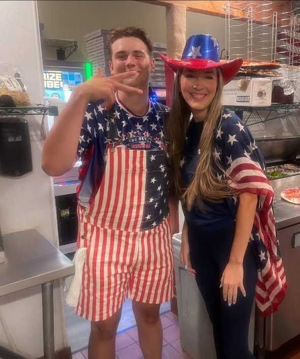 Two people in patriotic red, white, and blue outfits smiling indoors, one wearing a cowboy hat.