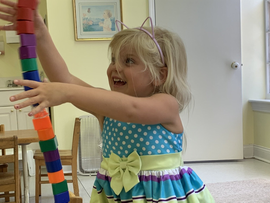 Child in a polka-dot dress reaching for a colorful stacking toy in a bright room
