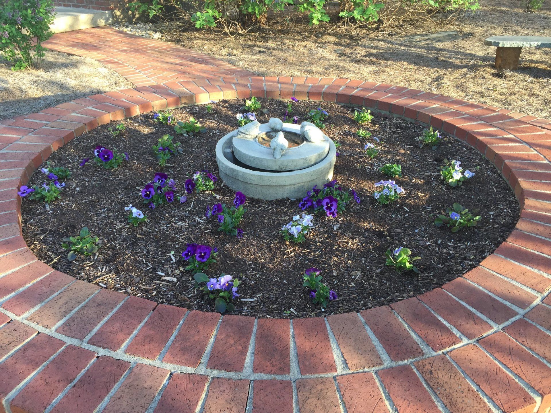 Brick-bordered circular flower bed with purple flowers and a small fountain in the center