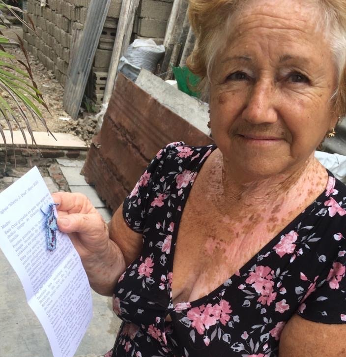 Woman holding a document outdoors beside a brick wall and sidewalk, wearing a black floral top, looking at the camera