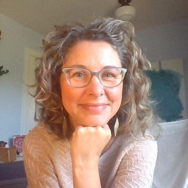 Smiling person with curly hair and glasses resting chin on hand indoors