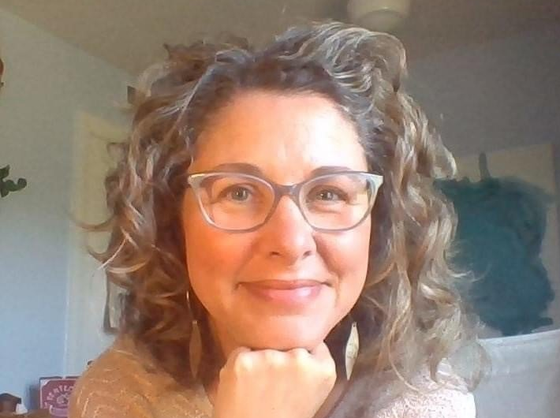 Woman with curly hair and glasses smiling indoors, resting her chin on her hand.