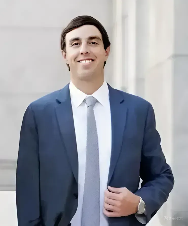 A professional headshot of a person in a blue suit, white shirt, and gray tie, smiling against an architectural background.