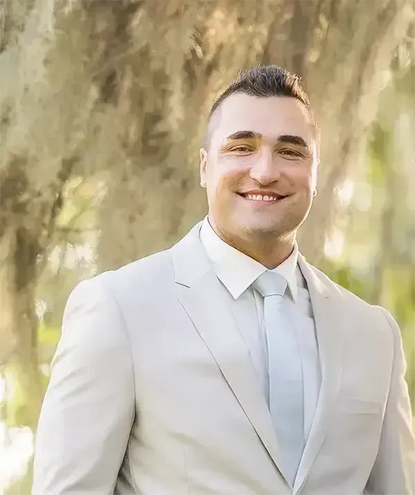 A smiling individual wearing a light gray suit and white tie, positioned against a soft-focus, golden outdoor background.