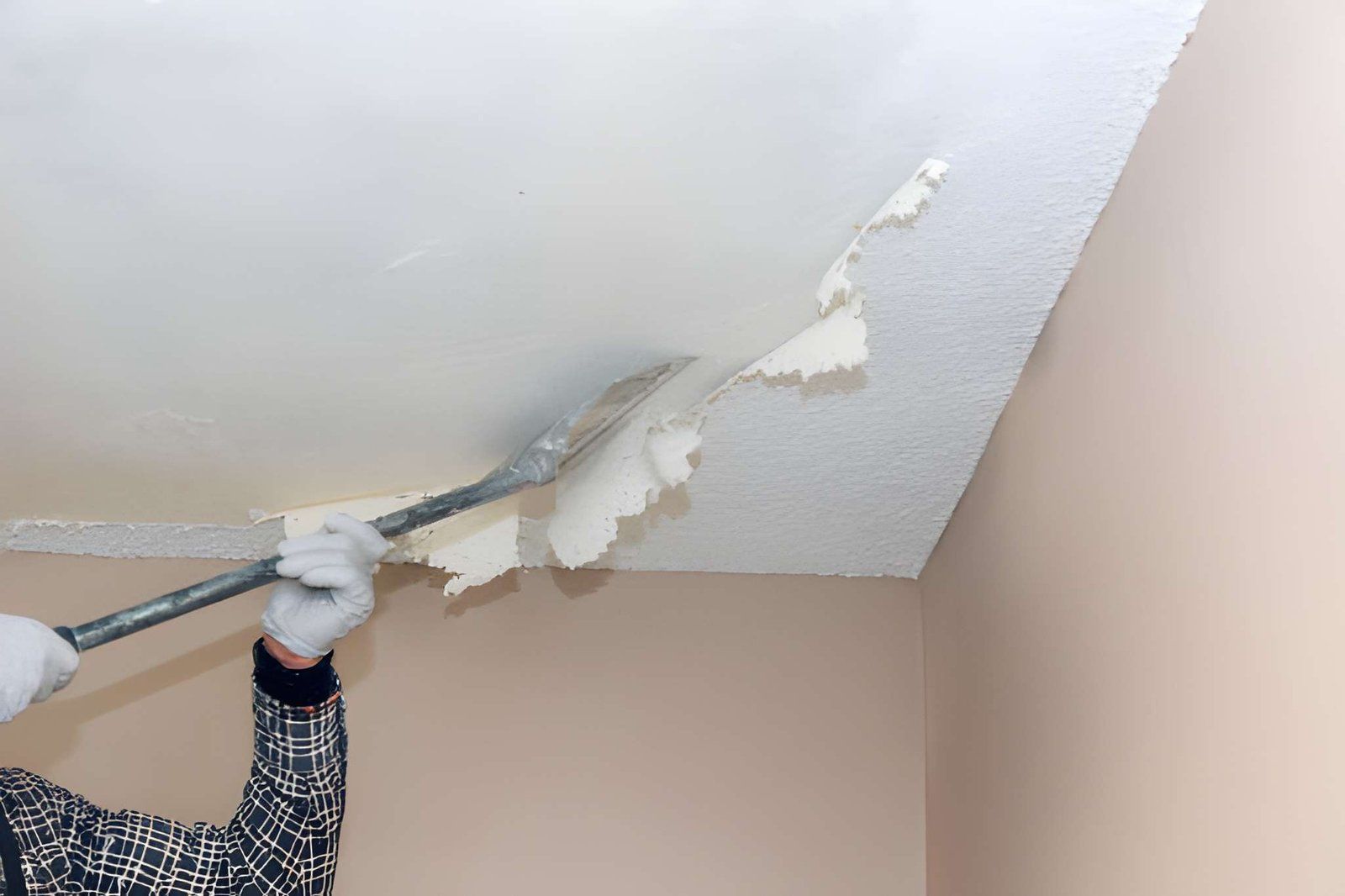 Person scraping peeling paint from a beige ceiling with a putty knife during indoor repair work