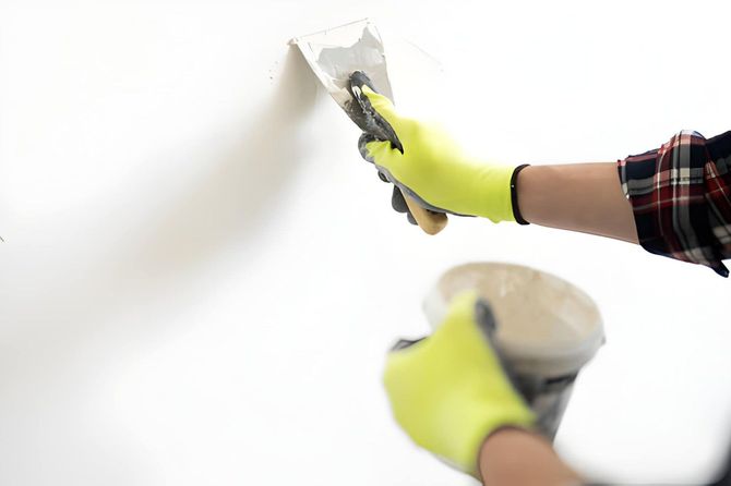 Hands in yellow gloves applying white paint to a wall with a putty knife and paint cup