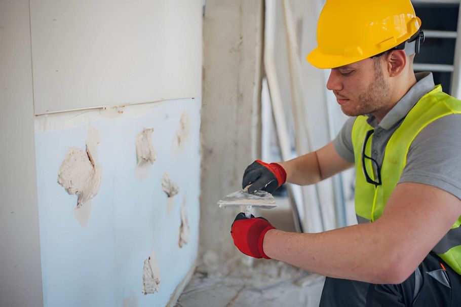 Worker in hard hat and safety vest scraping damaged wall plaster indoors