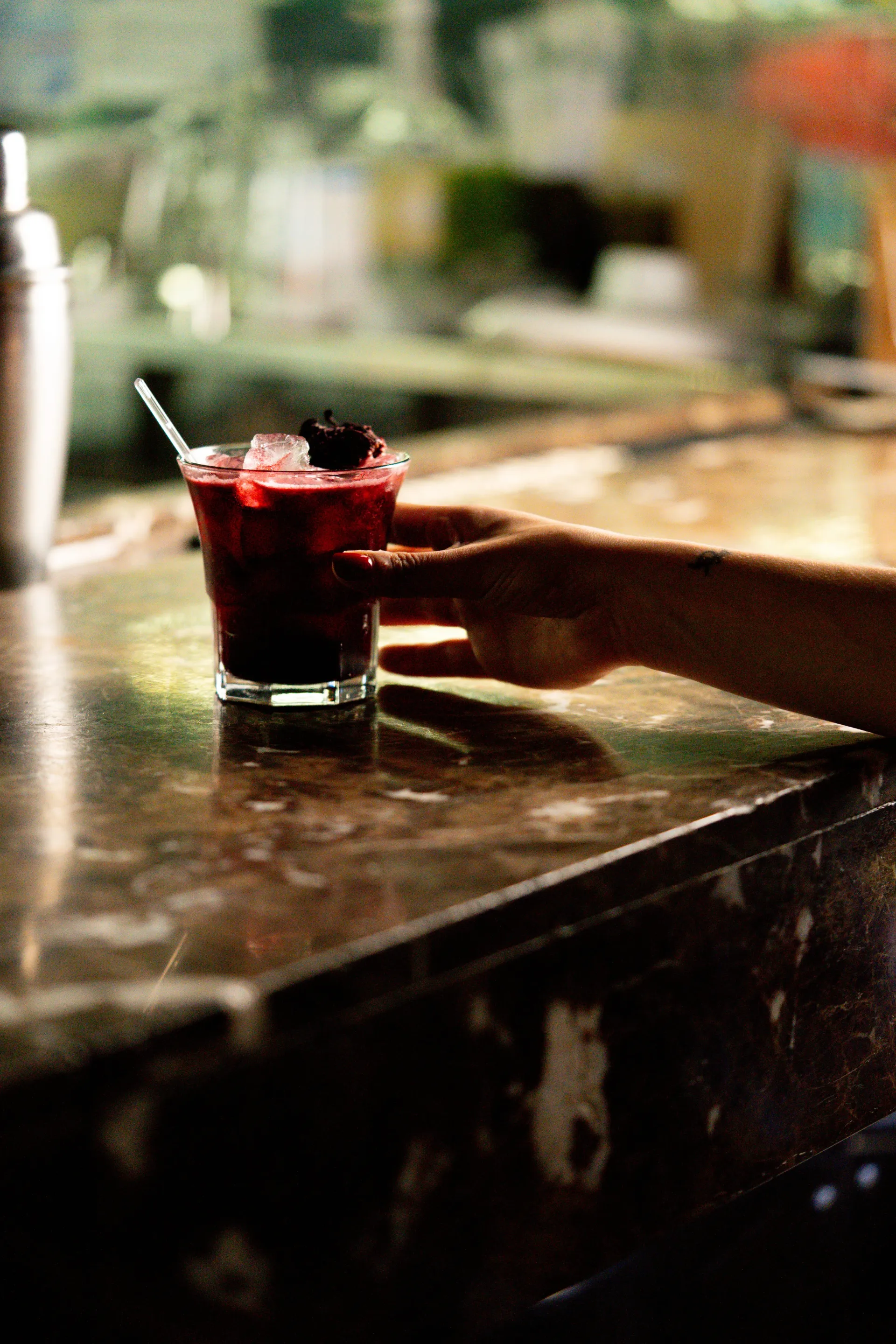 Una mano se extiende hacia una bebida roja con hielo y una pajita que está sobre la barra de un bar.