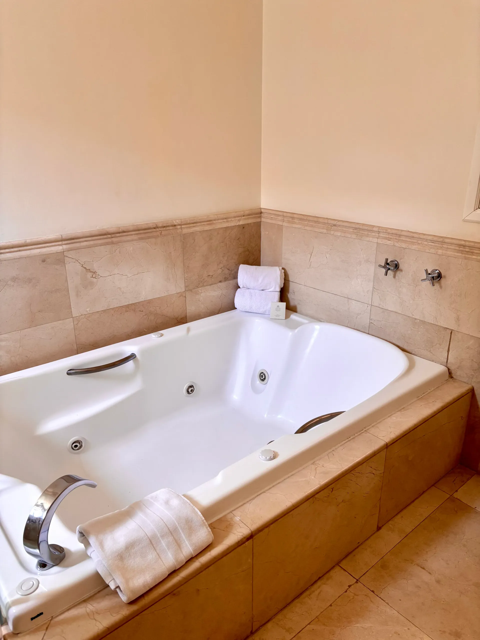 Baño de esquina con bañera de hidromasaje blanca, revestimiento de azulejos color canela y toallas dobladas en el borde.