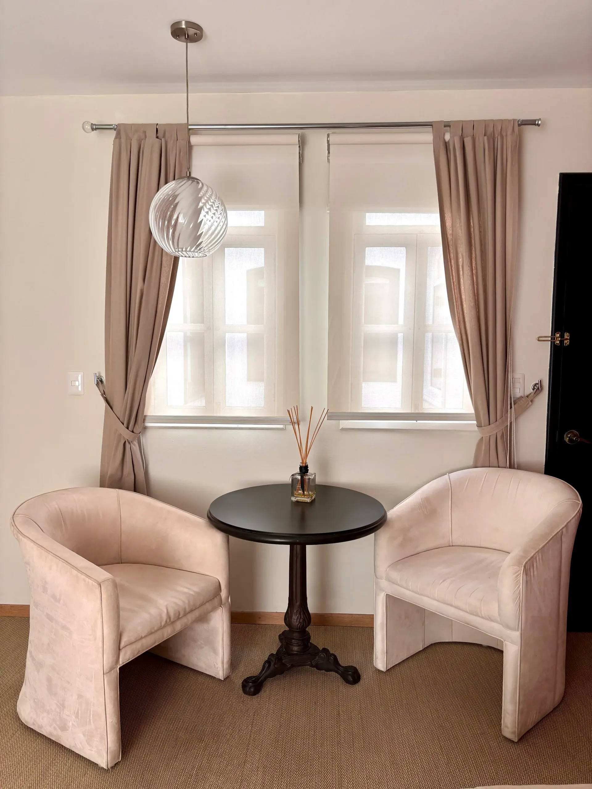 Dos sillones de color beige y una pequeña mesa redonda en una habitación luminosa junto a una ventana.