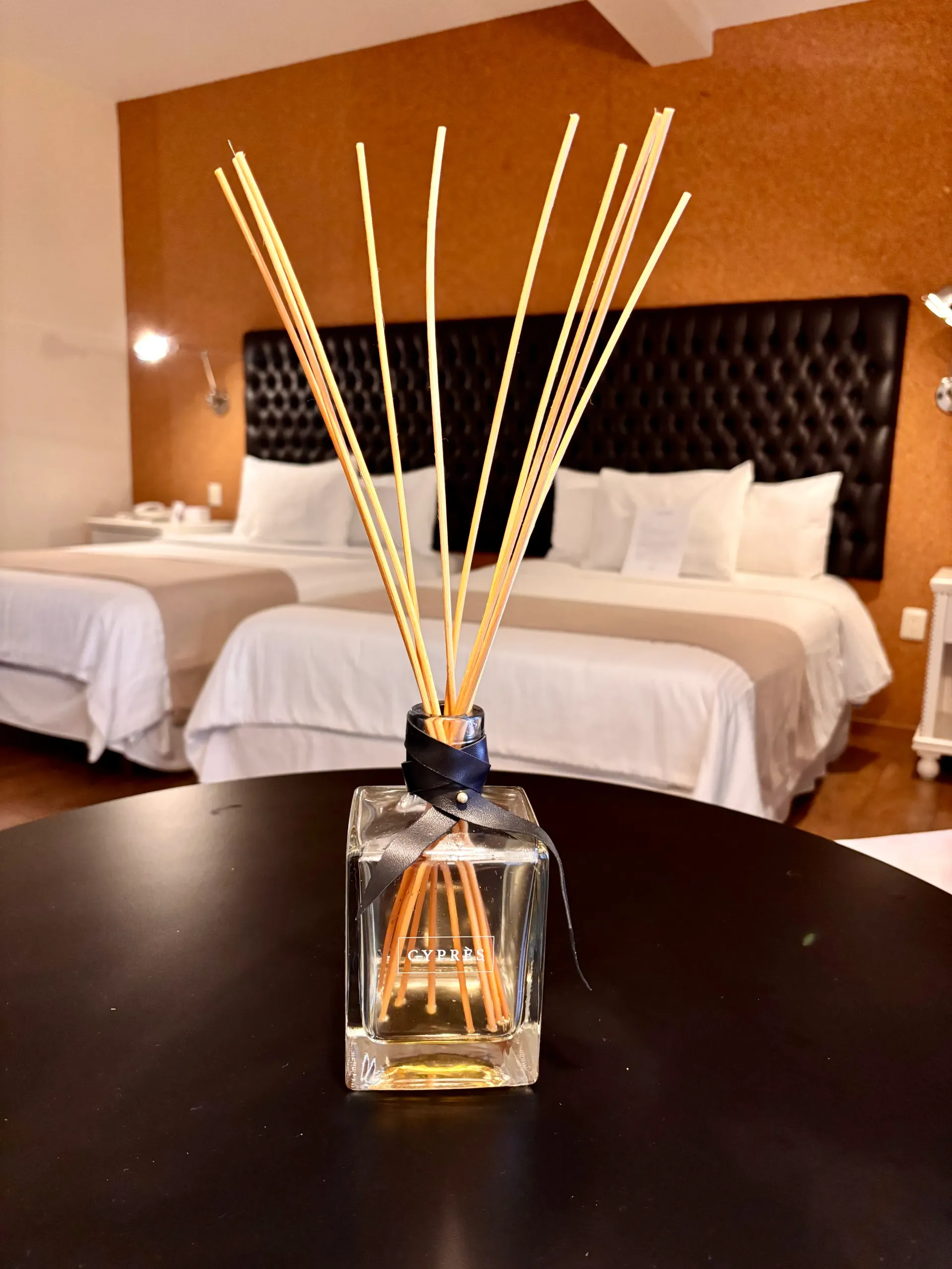 Difusor de varillas sobre una mesita de noche en una habitación de hotel de tonos cálidos con dos camas.