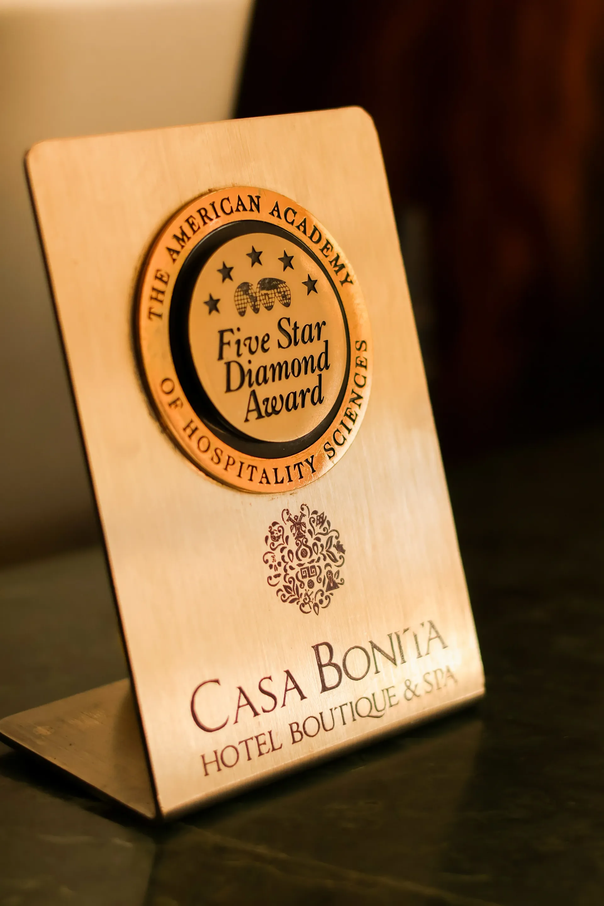 Placa de madera con un sello de premio redondo y el texto “Casa Bonita Hotel Boutique” sobre una mesa.