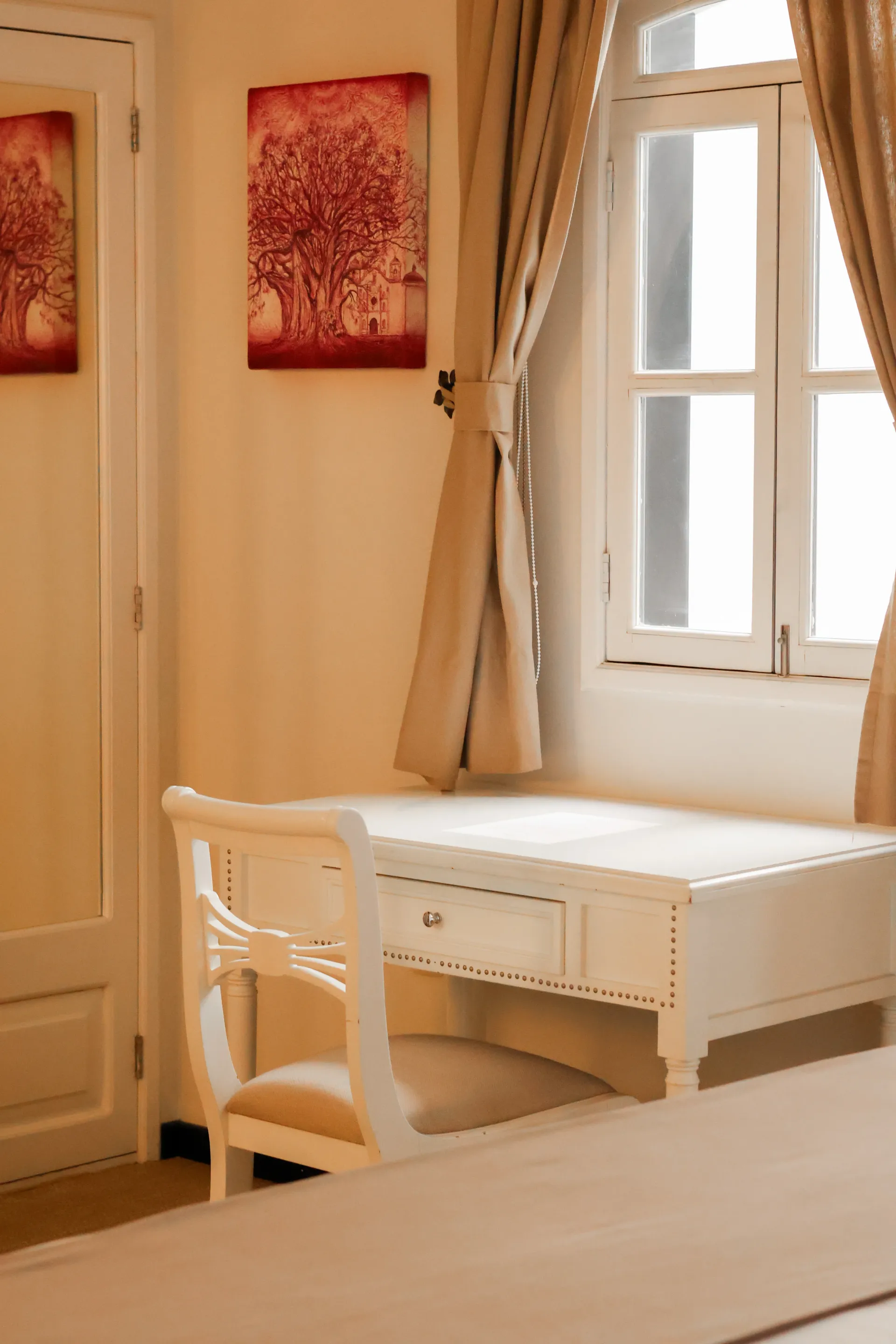 Escritorio y silla de color crema junto a una ventana soleada con cortinas beige.