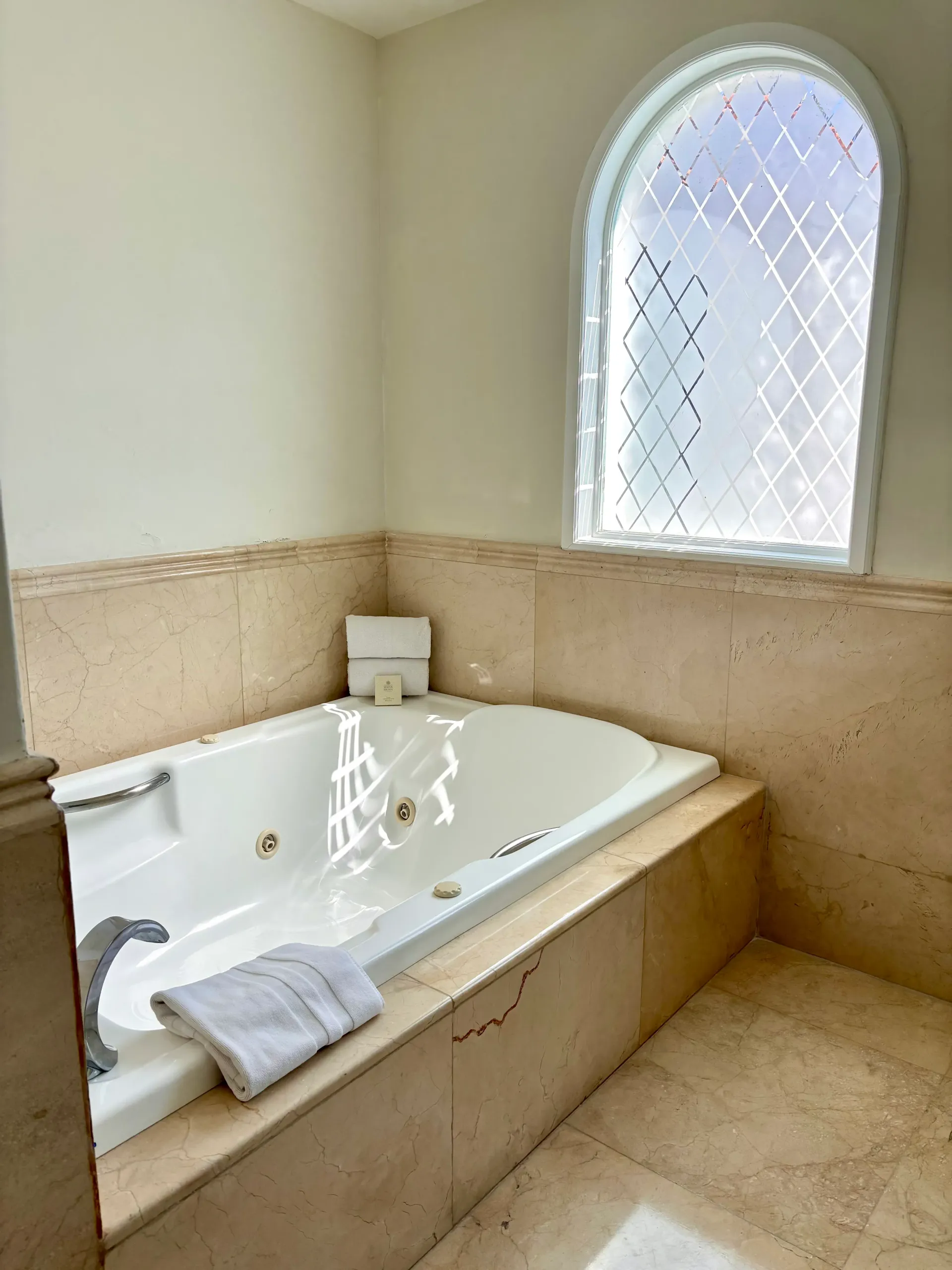 Un baño con azulejos beige, bañera, toalla y ventana arqueada esmerilada.