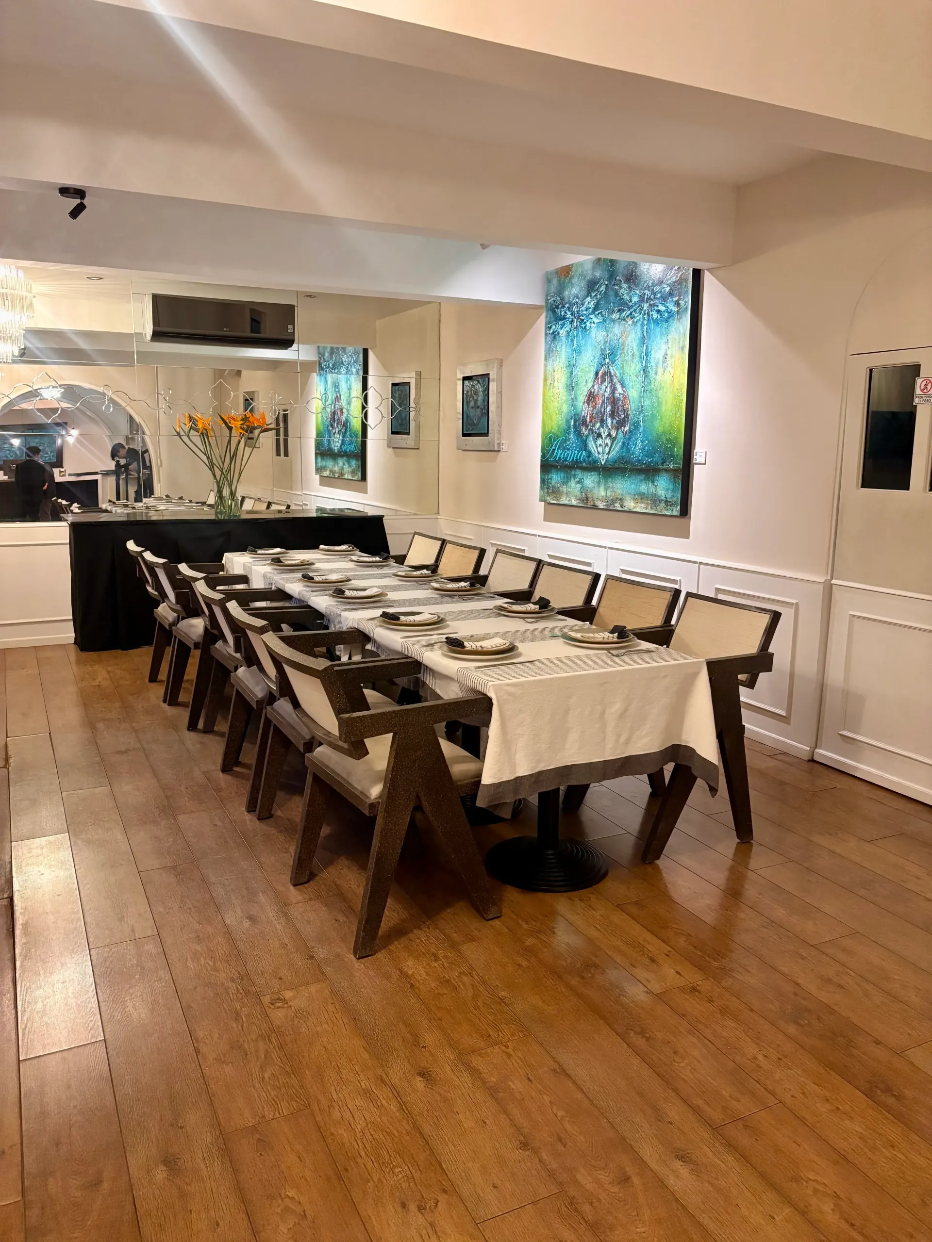 Mesa larga preparada para una comida en una habitación luminosa con coloridos cuadros en las paredes y suelos de madera.