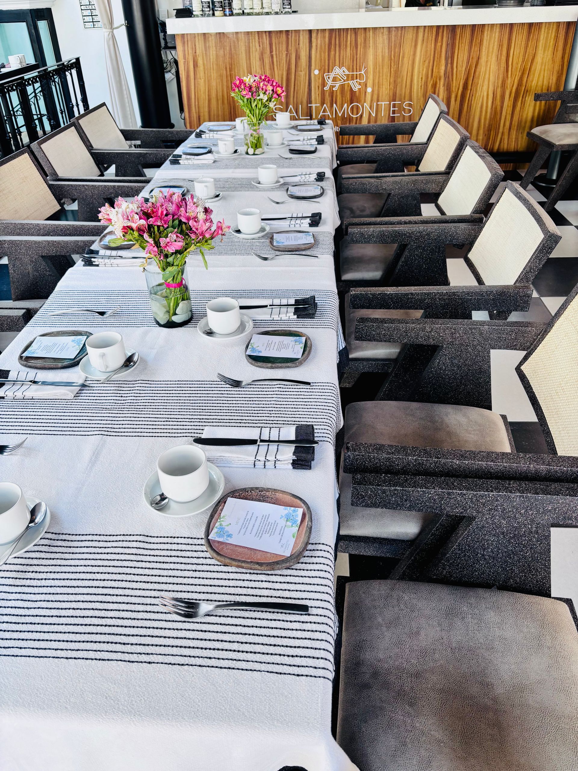 Mesa de larga para  con mantel de rayas blancas, centros de mesa florales y sillas acolchadas en un patio.