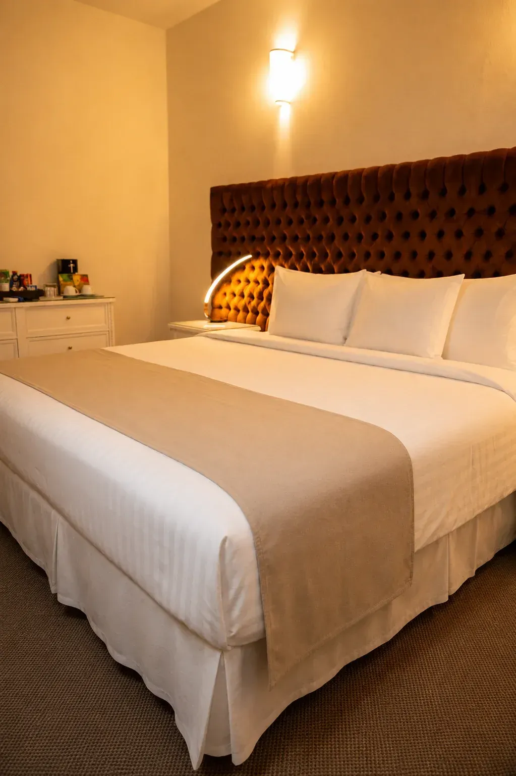 Acogedora habitación de hotel con una cama king size , almohadas blancas, manta beige e iluminación cálida en las paredes.