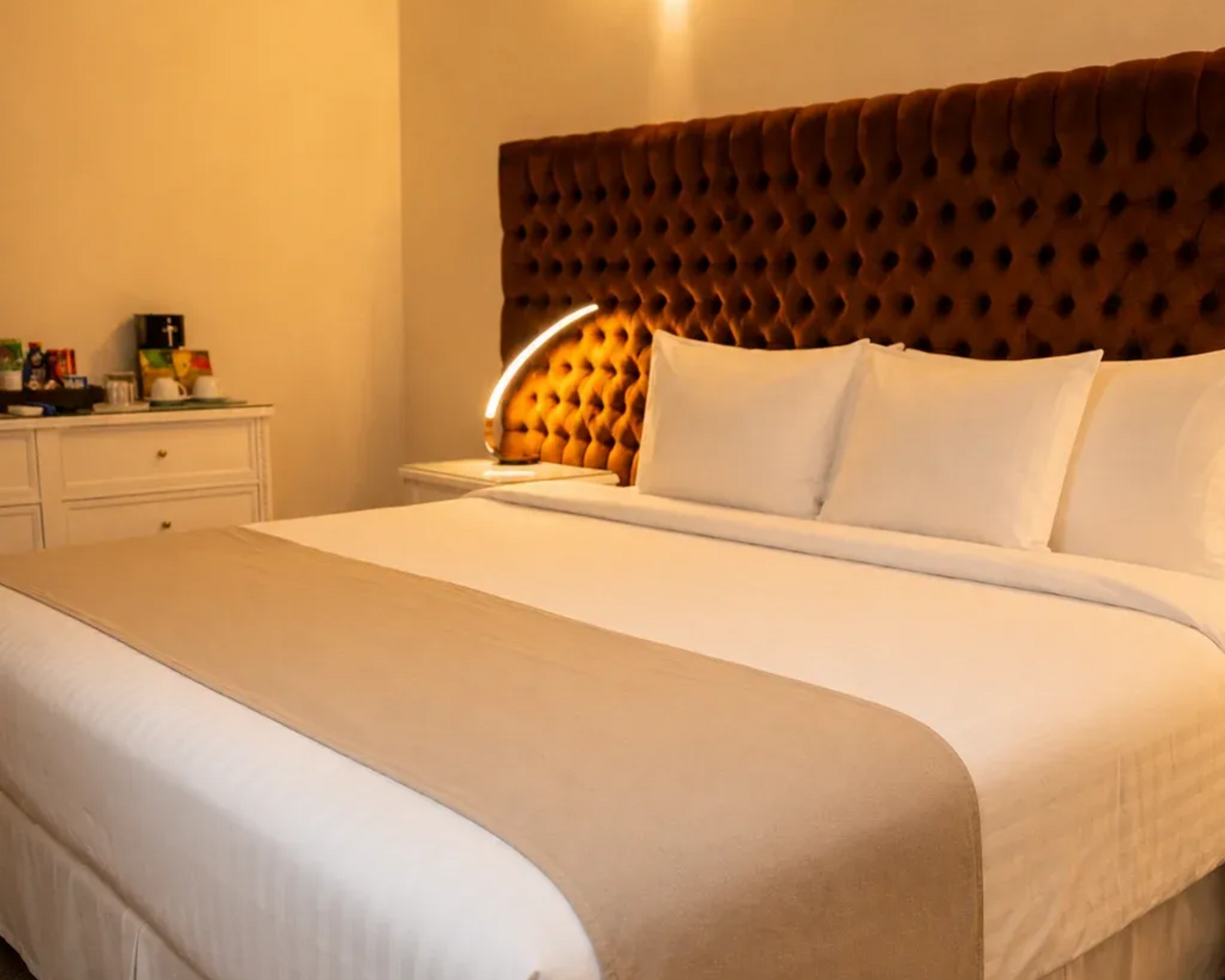 Cama con ropa de cama blanca, camino de mesa beige y cabecero capitoné marrón en un dormitorio con iluminación cálida.