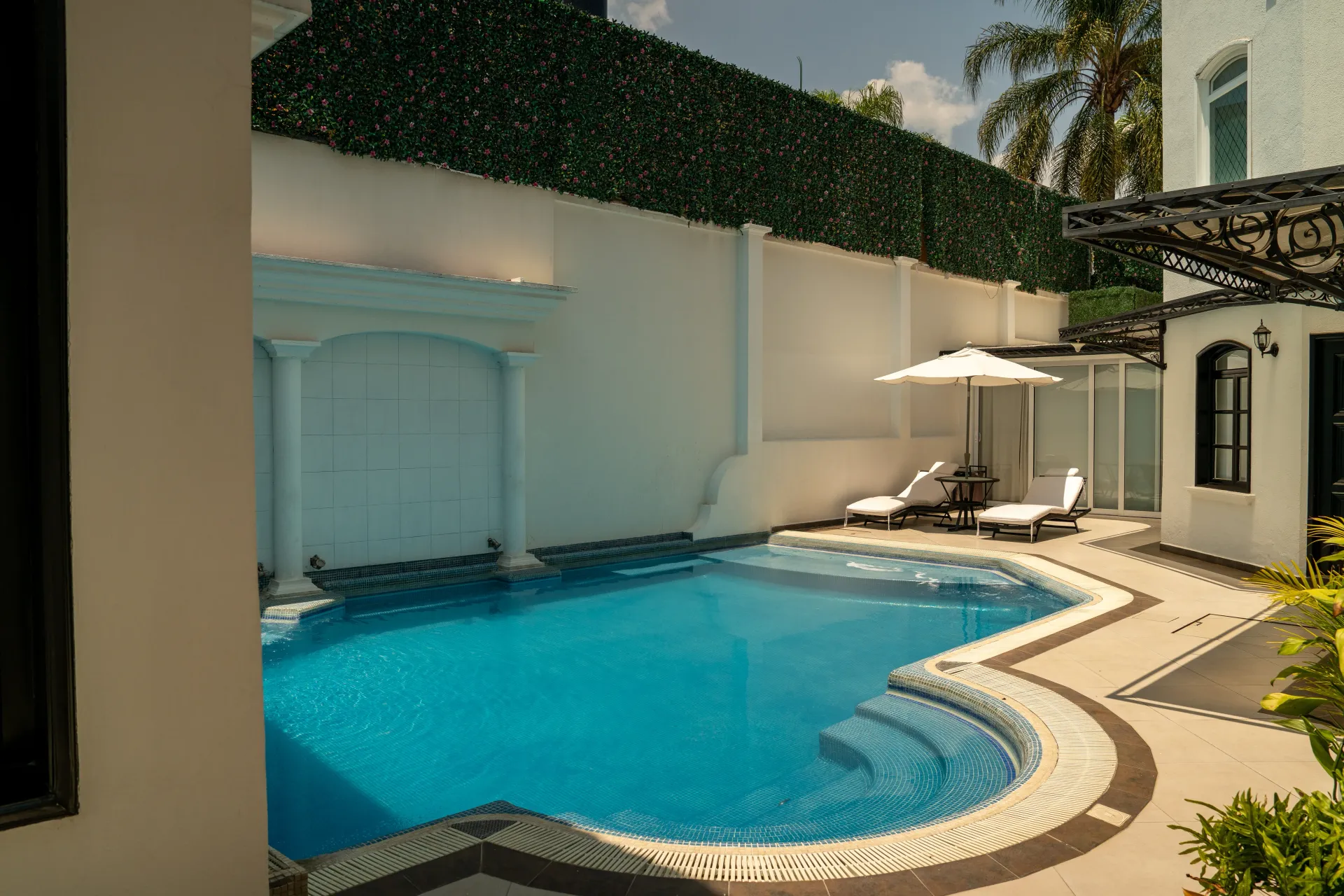 Piscina moderna al aire libre con agua azul, tumbonas y sombrillas blancas junto a un patio embaldosado.