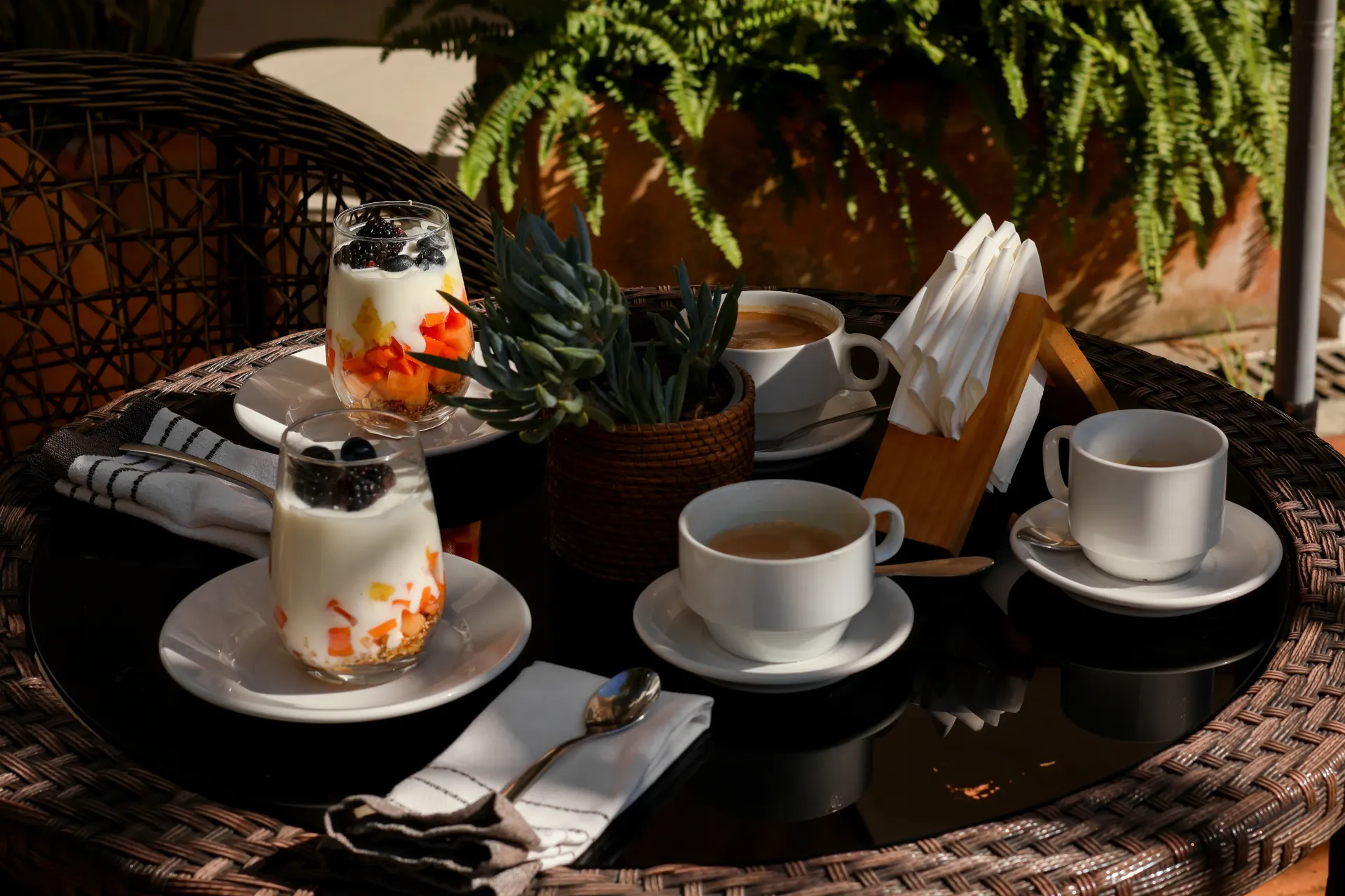 Tazas de café y copas de postre sobre una mesa de mimbre en el patio, con una bandeja negra y un helecho al fondo.