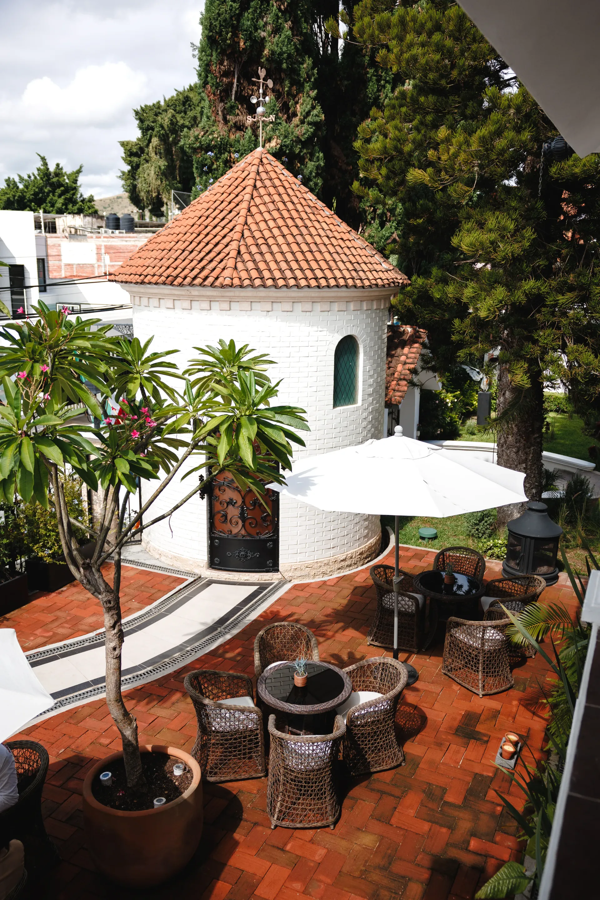 Casita blanca con tejado de tejas rojas y patio ajardinado, con plantas en macetas, una mesa y una sombrilla blanca.