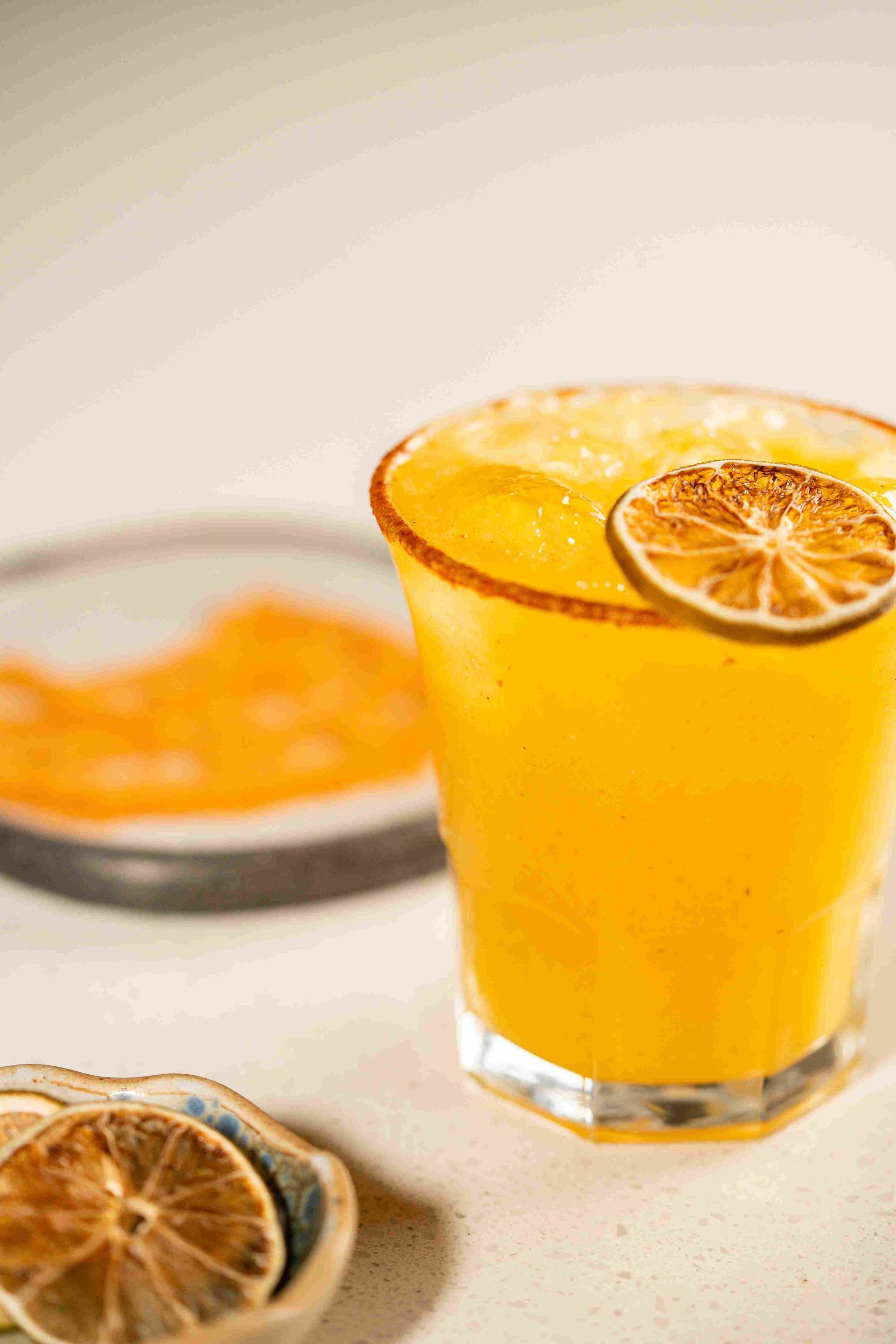 Vaso de bebida de naranja con una rodaja de cítrico, junto a una bandeja de fruta borrosa sobre una mesa clara.