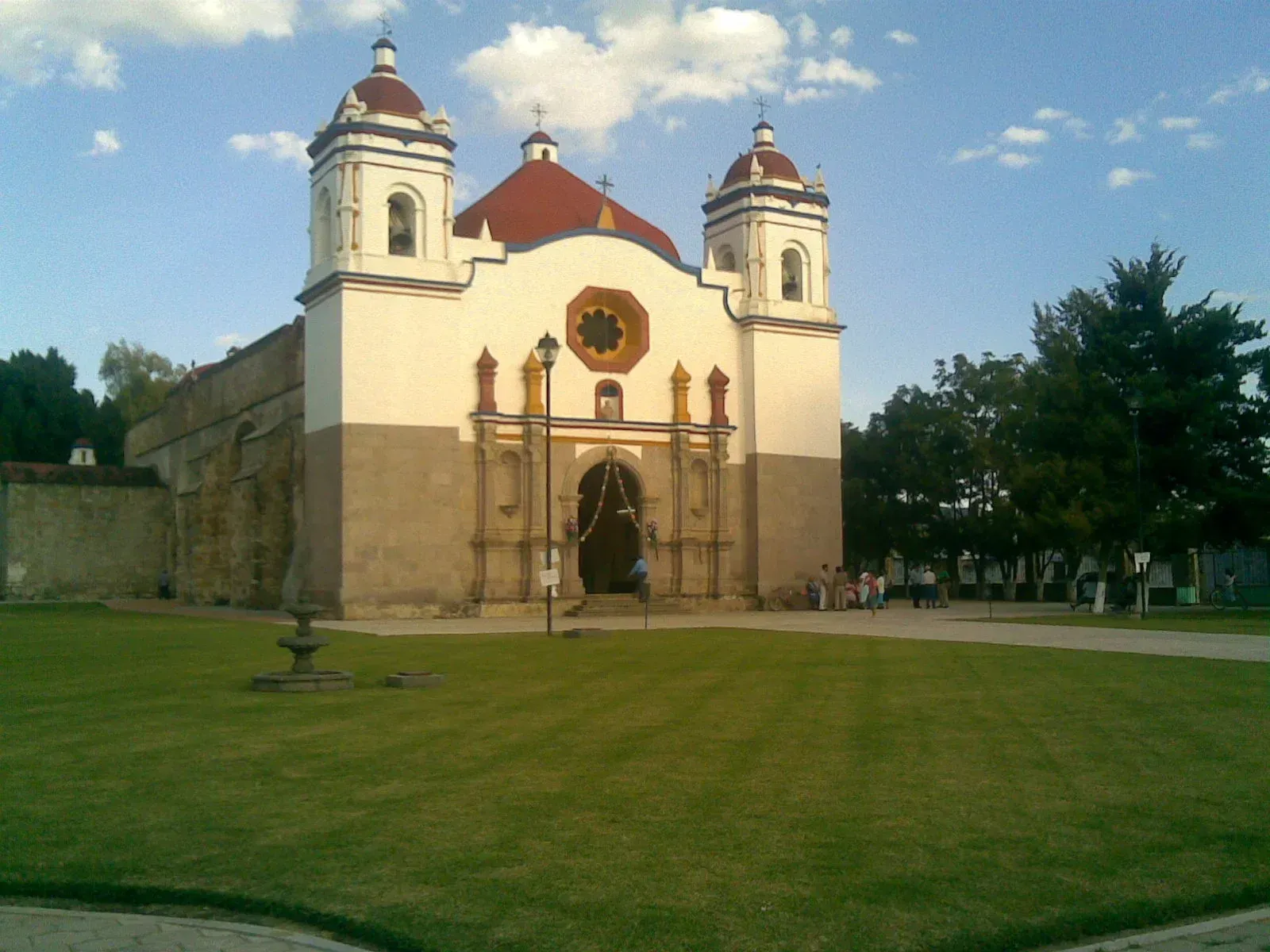 Iglesia histórica con dos torres blancas y techo rojo junto a un gran césped verde bajo un cielo parcialmente nublado.