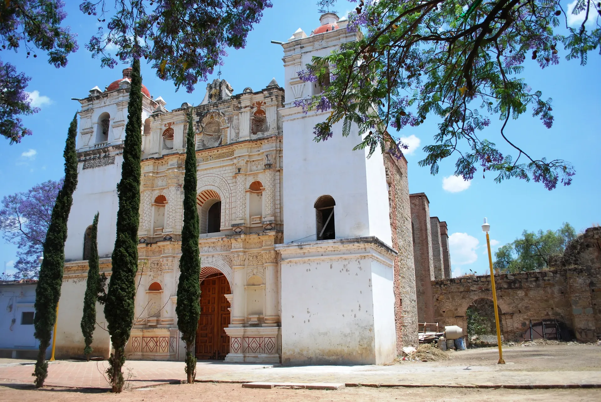 Antigua iglesia de piedra blanca con cipreses y cielo azul, vista desde una calle soleada.