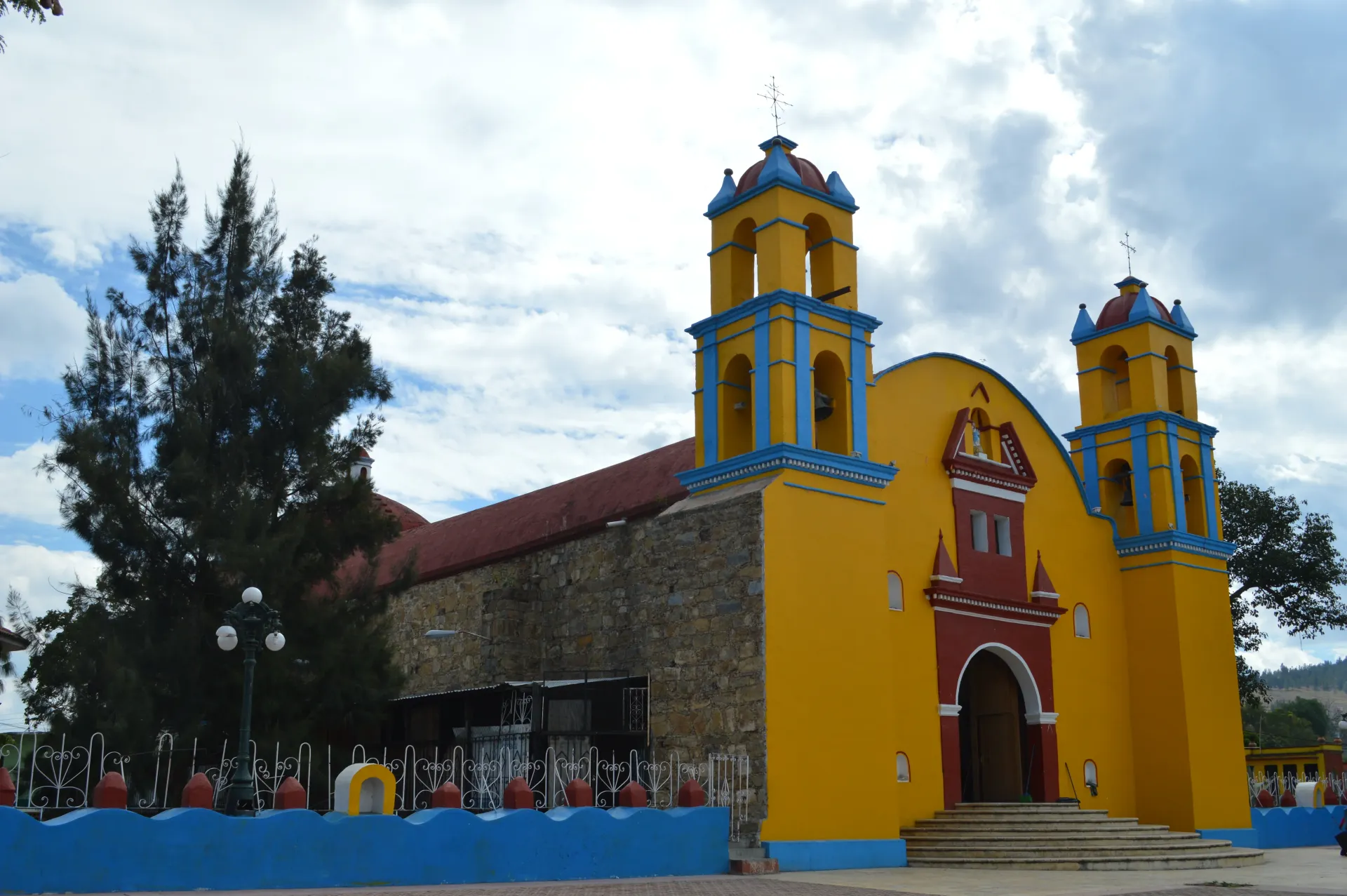 Iglesia amarilla con torres azules y techo rojo junto a un muro azul bajo un cielo nublado.