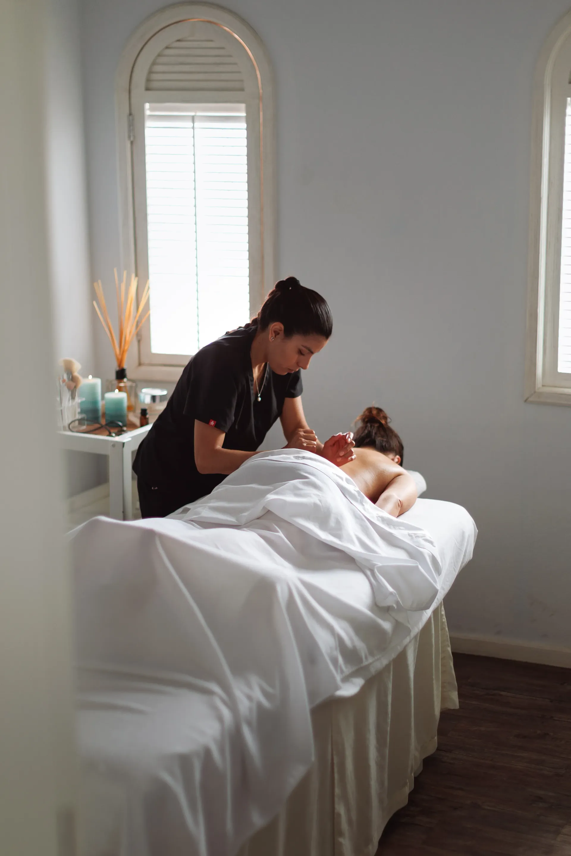 Terapeuta de spa realizando un tratamiento facial a una clienta en una camilla blanca en una habitación luminosa.