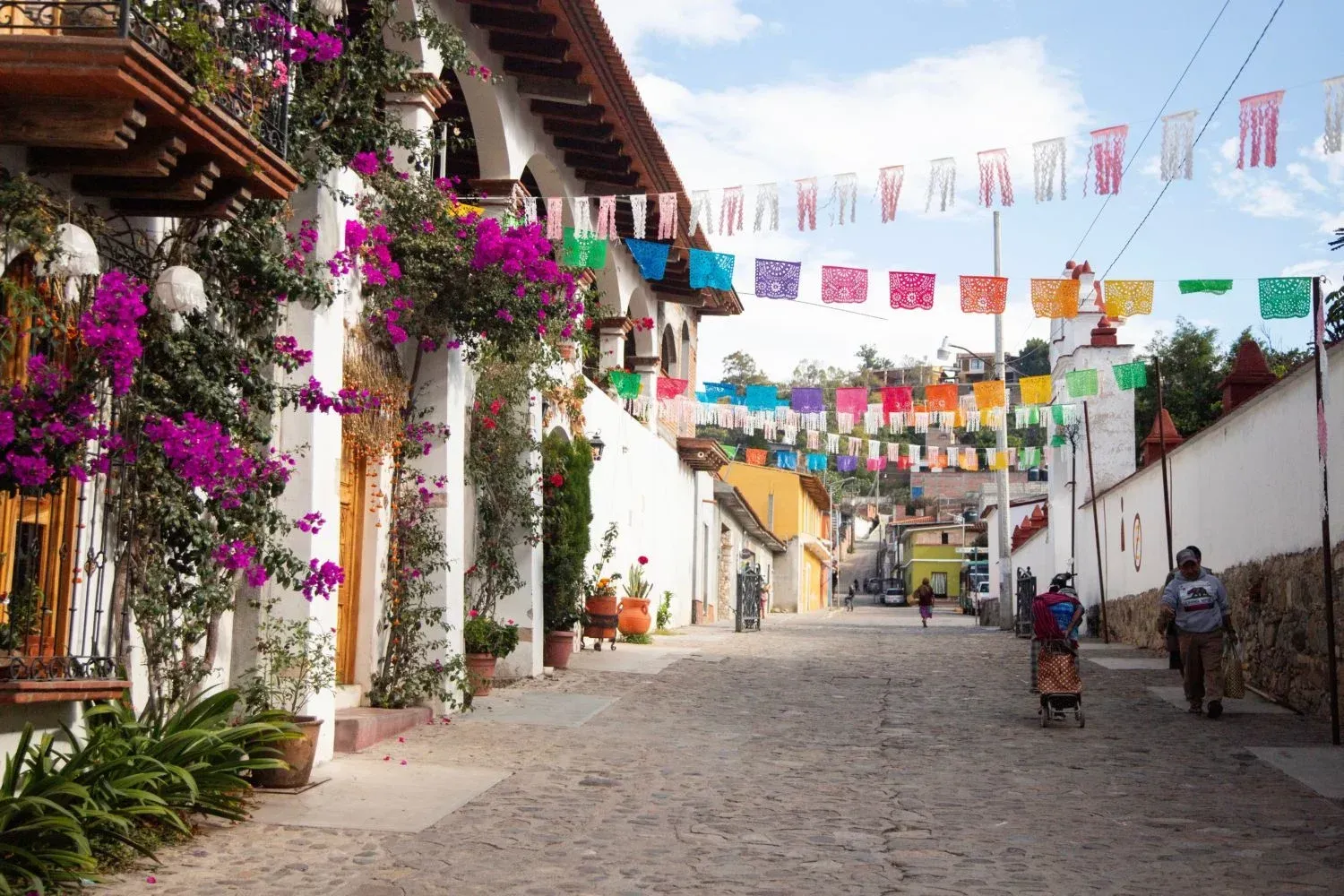 Calle empedrada con coloridas banderas, edificios blancos y flores moradas bajo un cielo brillante.