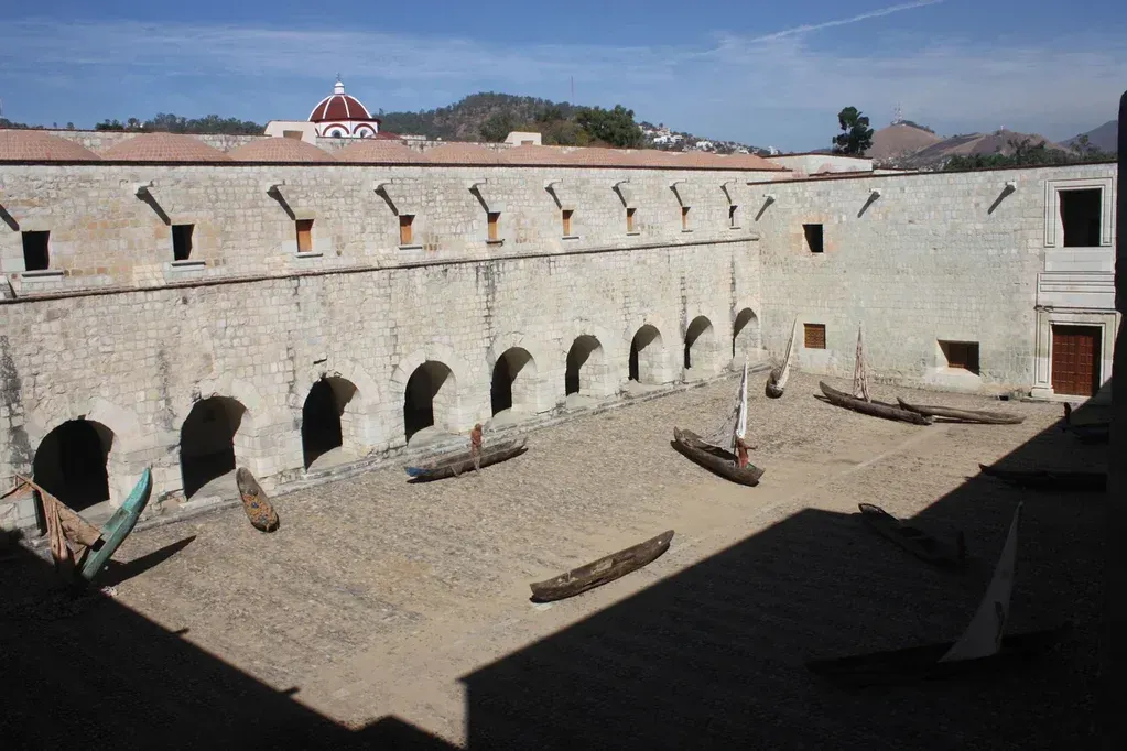 Patio histórico de piedra con muros arqueados y una torre de techo rojo bajo un cielo azul.
