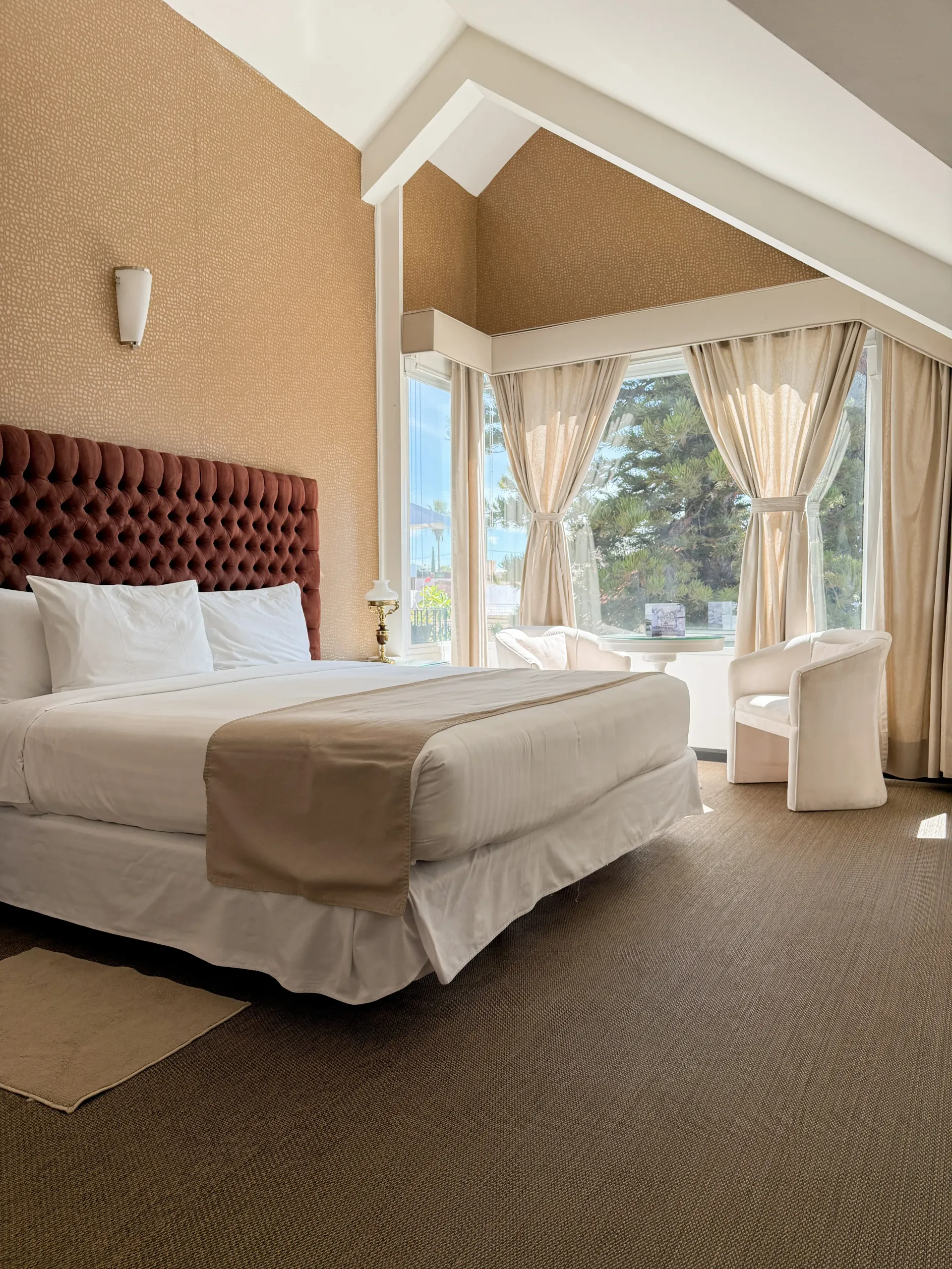 Habitación de hotel moderna con cama king size, cabecero capitoné y grandes ventanales con cortinas y sillas.