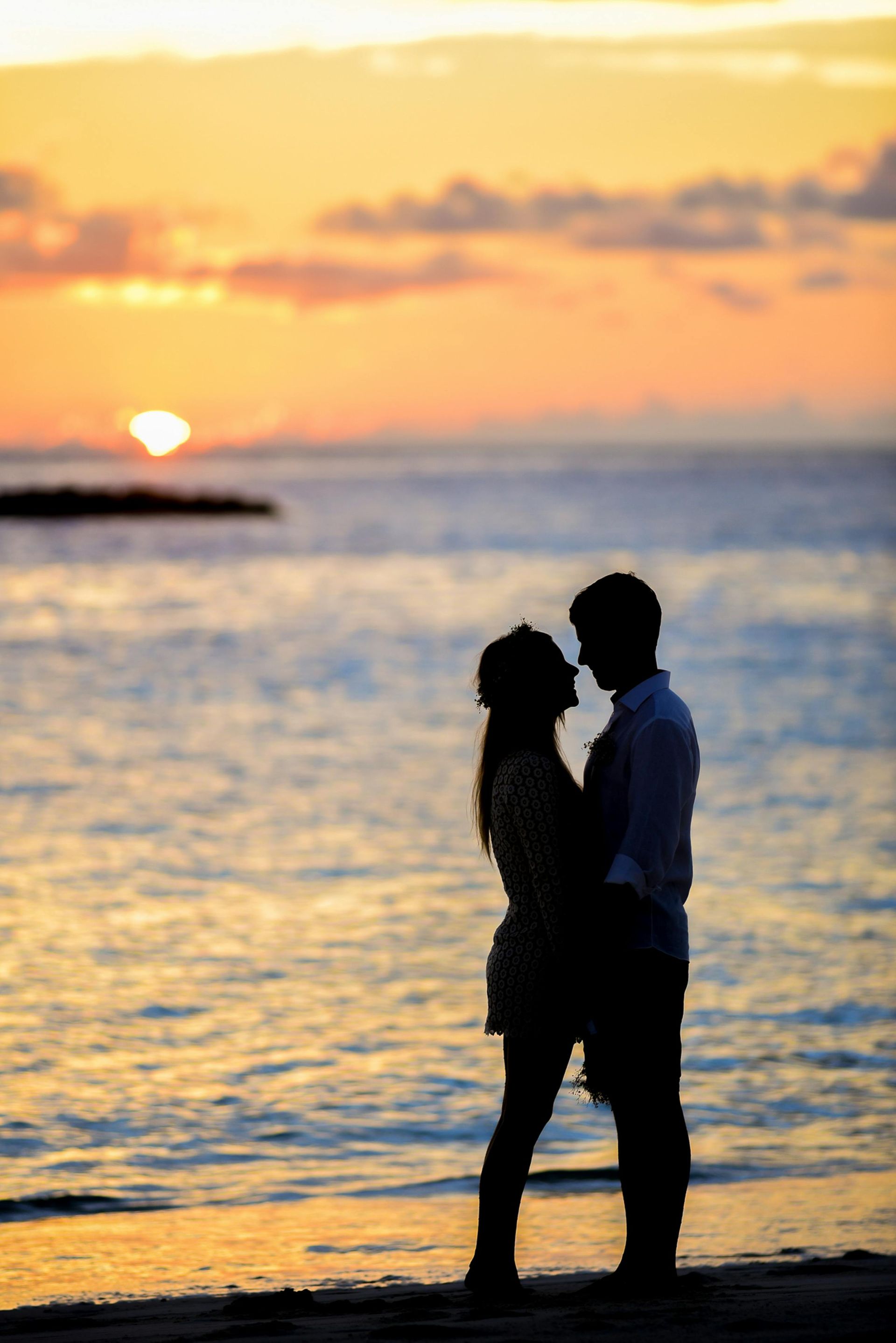 Pareja recortada contra el agua abrazándose al atardecer, con cielo anaranjado y mar en calma.
