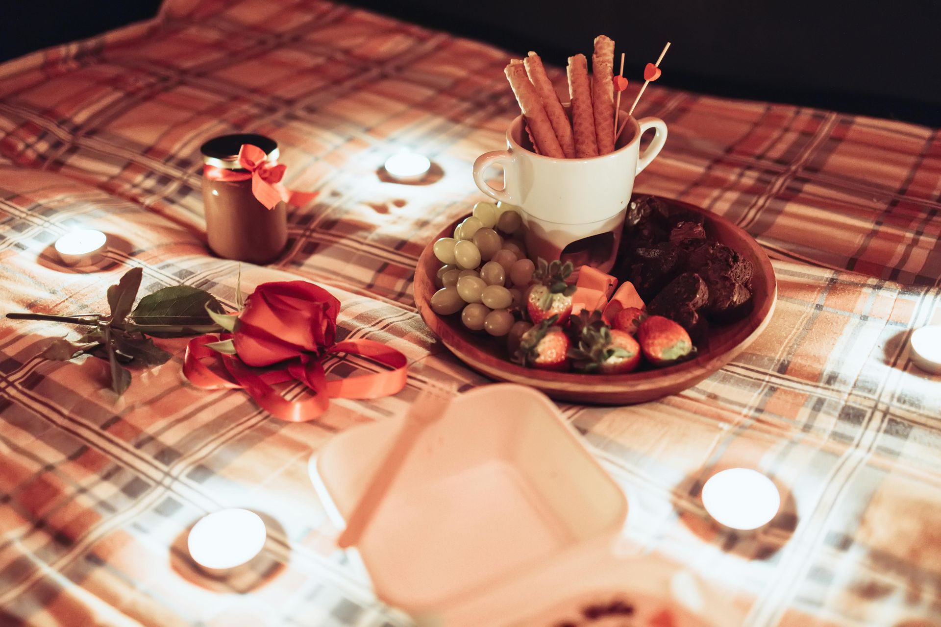 Mesa de picnic iluminada con velas con un plato de uvas y bocadillos, rosas y una taza de galletas sobre una manta a cuadros.