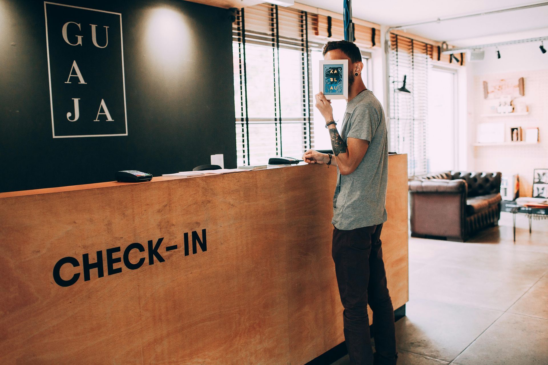 Mostrador de recepción de hotel con una persona usando un teléfono inteligente, mostrador de madera y zona de estar al fondo.
