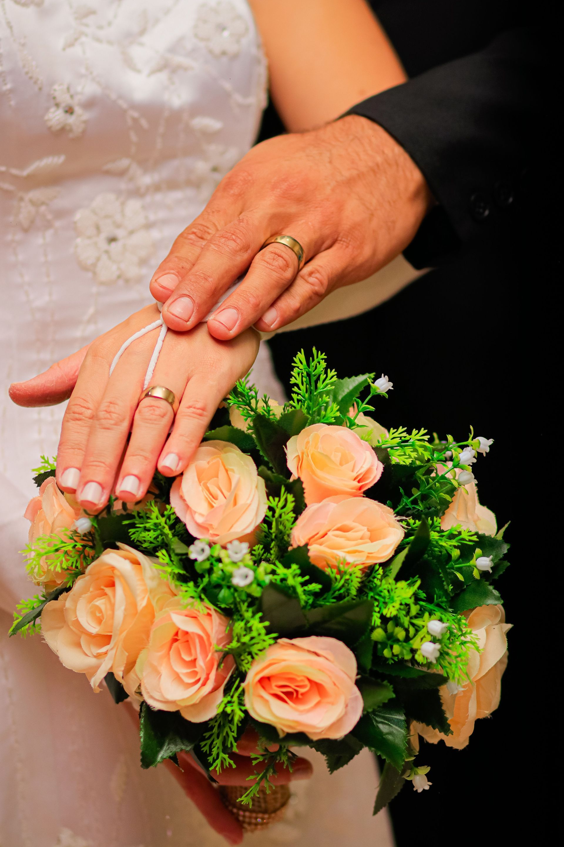 Manos con anillos de boda descansando sobre un ramo de rosas color melocotón.