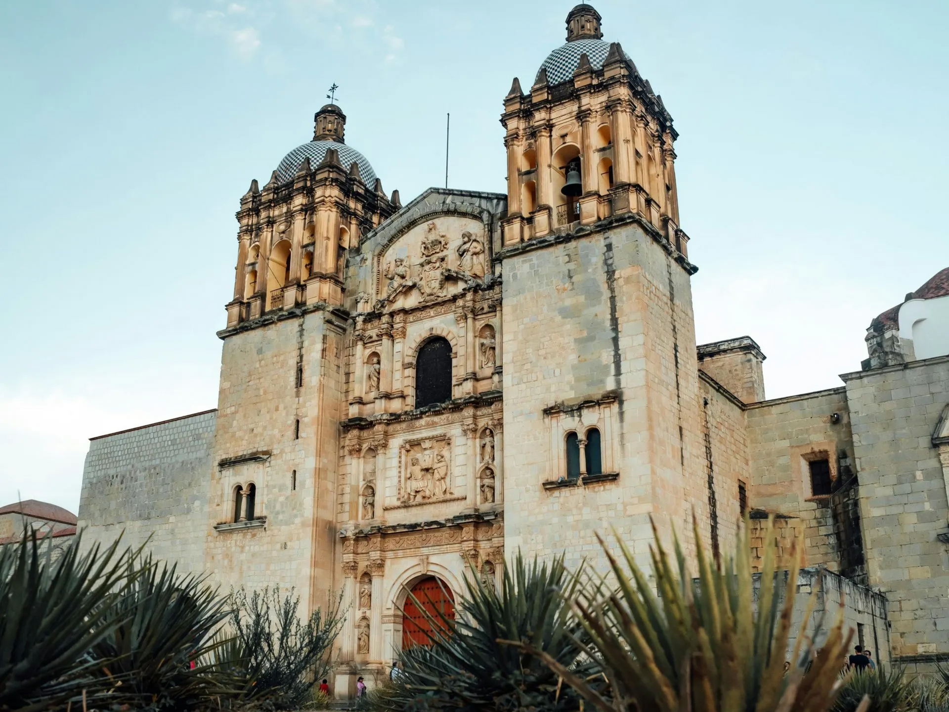 Iglesia histórica de piedra con dos campanarios y una puerta arqueada roja contra un cielo azul.