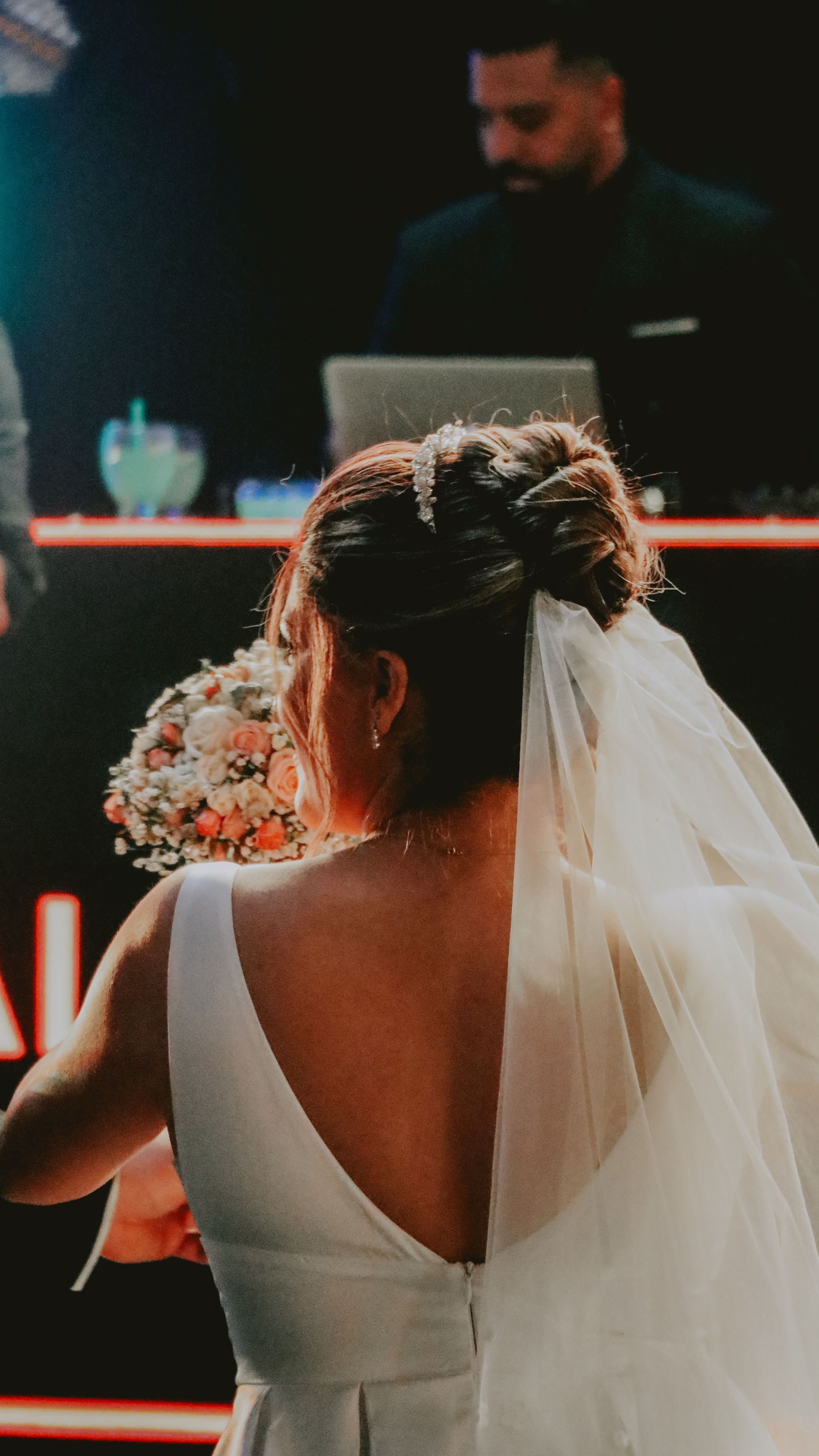 La novia, vestida de blanco y con velo, sentada frente a un orador en una mesa de ceremonia decorada.