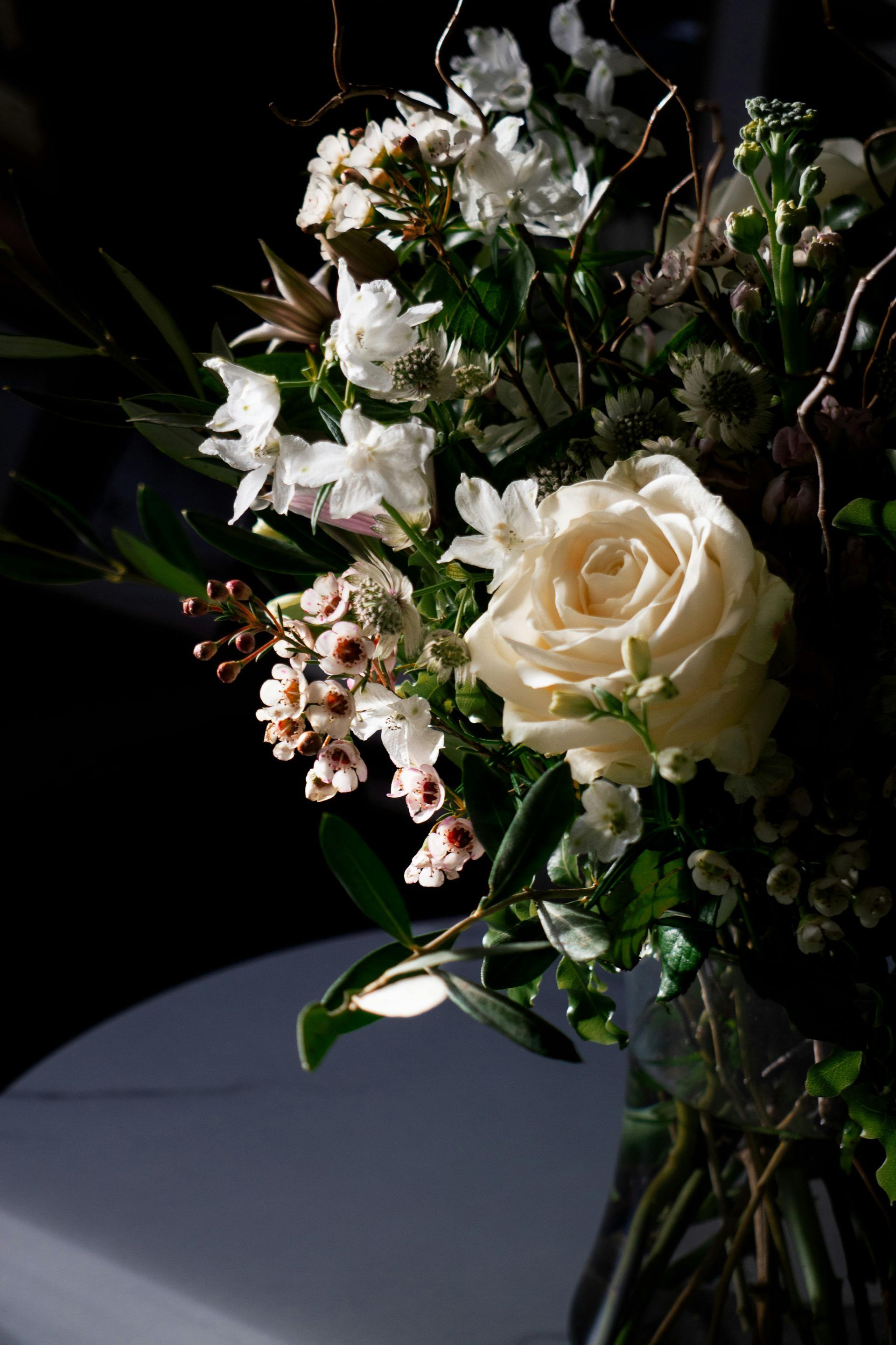 Ramo floral en tonos crema y blanco con rosas y follaje en un jarrón oscuro sobre un fondo negro.