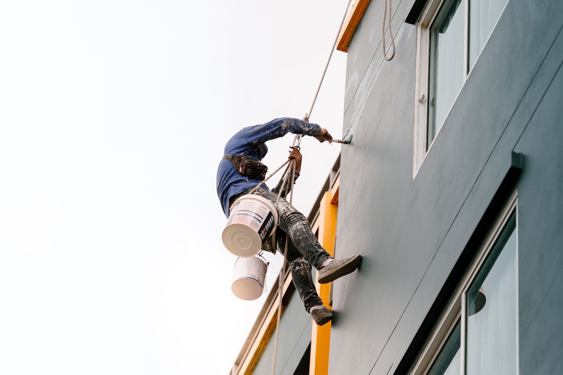 Painter suspended on ropes, painting a teal building exterior; white paint bucket.