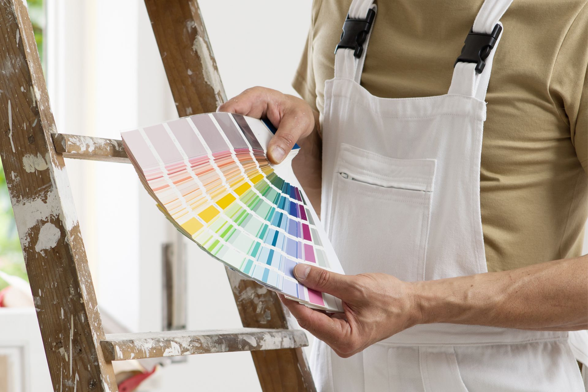 Person in paint overalls holding a color swatch by a wooden ladder.