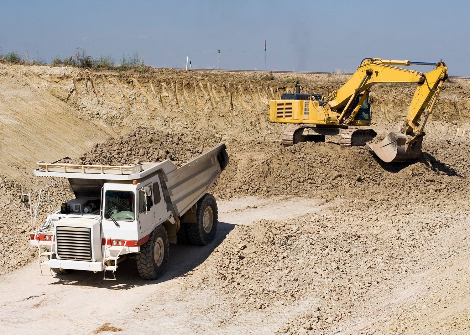 A yellow excavator loads dirt into a white dump truck at a construction site. A yellow excavator loads dirt into a white dump truck at a construction site.