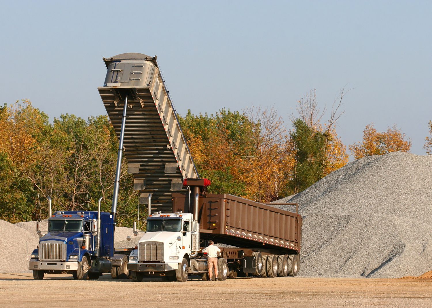 Two dump trucks unloading gravel at a quarry; a man stands near. Two dump trucks unloading gravel at a quarry; a man stands near.