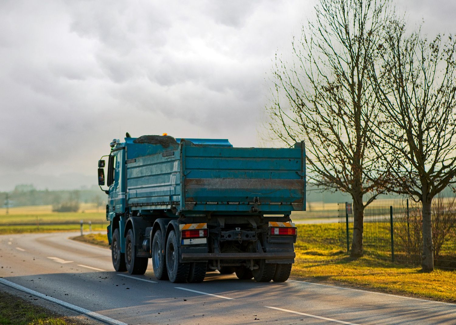 Blue dump truck on a road, driving away, with fields and trees in the background under a cloudy sky. Blue dump truck on a road, driving away, with fields and trees in the background under a cloudy sky.